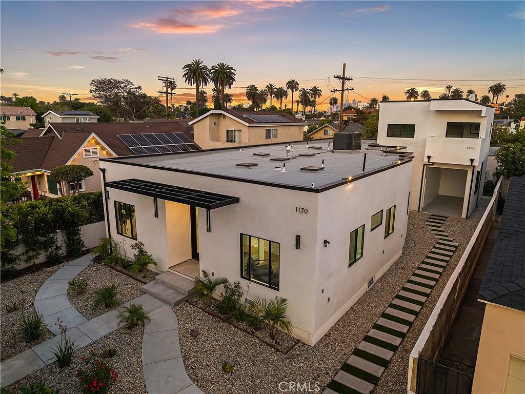 Aerial view of 1170 S Lucerne Blvd at twilight, a modernist flat-roof residence glowing under a pink Los Angeles sunset with palm trees on the horizon.